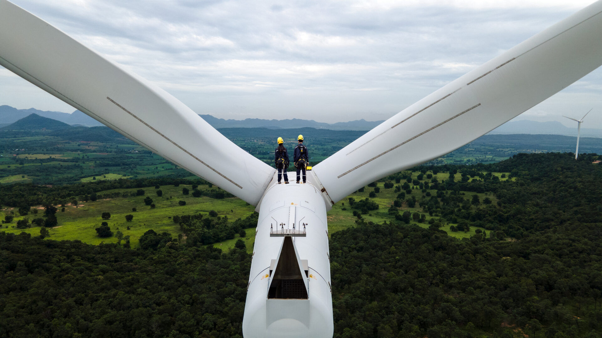 Two Electric engineer wearing Personal protective equipment working on top of wind turbine farm.